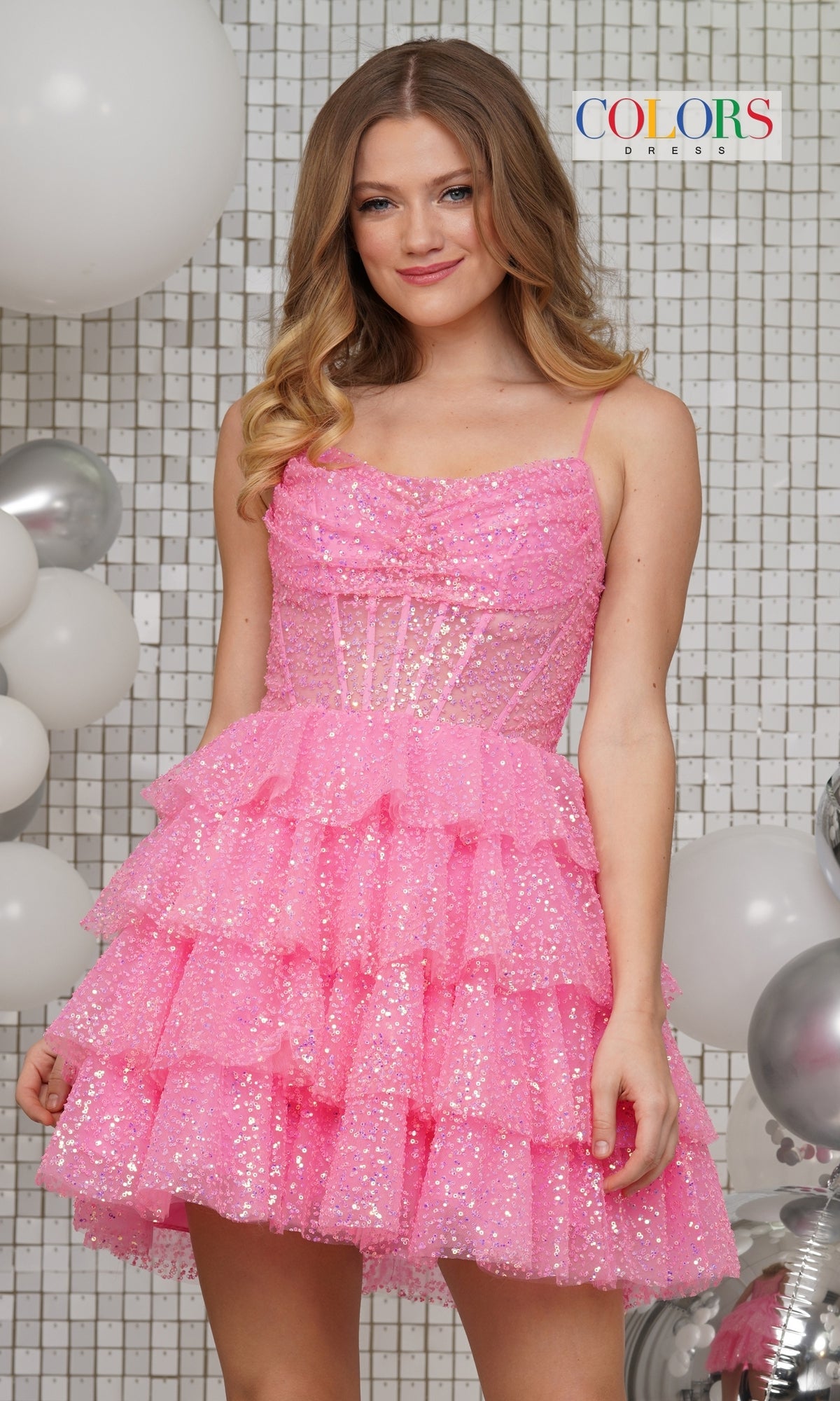 A young woman wears the Colors Dress Sequin Tulle Homecoming Dress 3345, featuring pink tiers and a sleeveless design, as she stands before a tiled wall with white and silver balloons.